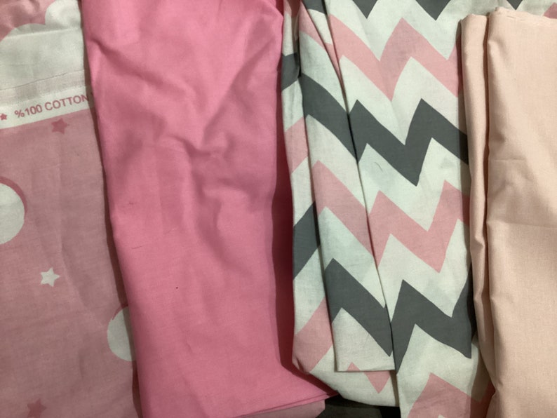 K&ouml;nnte beinhalten: Auswahl an gefalteten Stoffst&uuml;cken in Rosa-, Wei&szlig;- und Graut&ouml;nen. Ein St&uuml;ck hat ein Zickzackmuster, ein anderes einen Stern- und Monddruck. Der Text "100% COTTON" ist auf einem der Stoffe zu sehen.