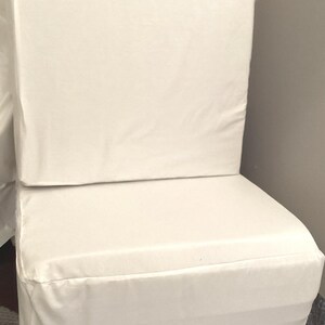 Puede incluir: Un cojín de silla rectangular blanco con un asiento y respaldo gruesos y acolchados. El cojín está hecho de una tela suave y lisa y tiene un diseño simple y moderno.