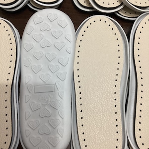 May include: A close-up of several white leather shoe insoles. Some insoles have a heart pattern on the bottom, while others have a dotted pattern.