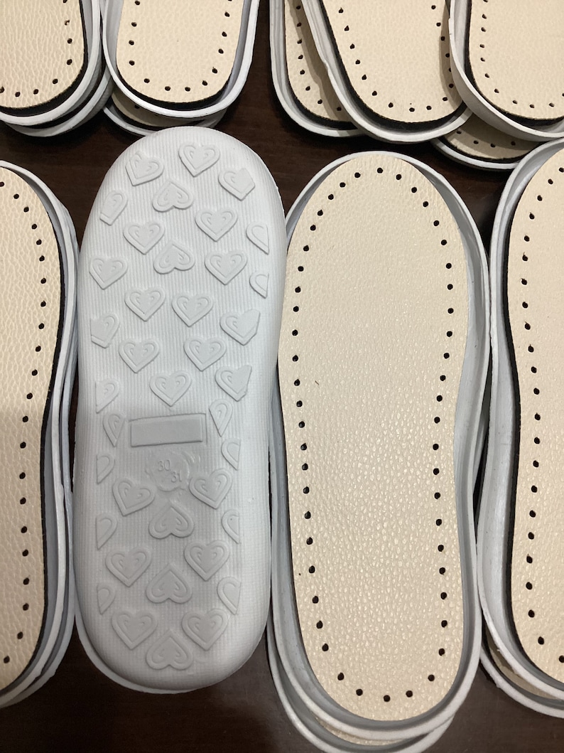 May include: A close-up of several white leather shoe insoles. Some insoles have a heart pattern on the bottom, while others have a dotted pattern.