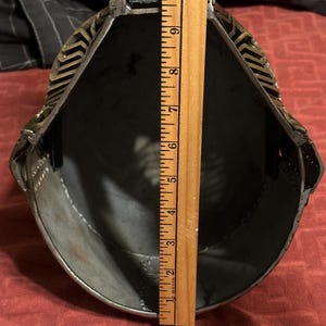 May include: A metal helmet viewed from the interior, featuring a dark grey inner surface and a decorative brass-toned exterior trim. A wooden ruler is placed vertically across the opening, showing a measurement of approximately 10 inches in length.