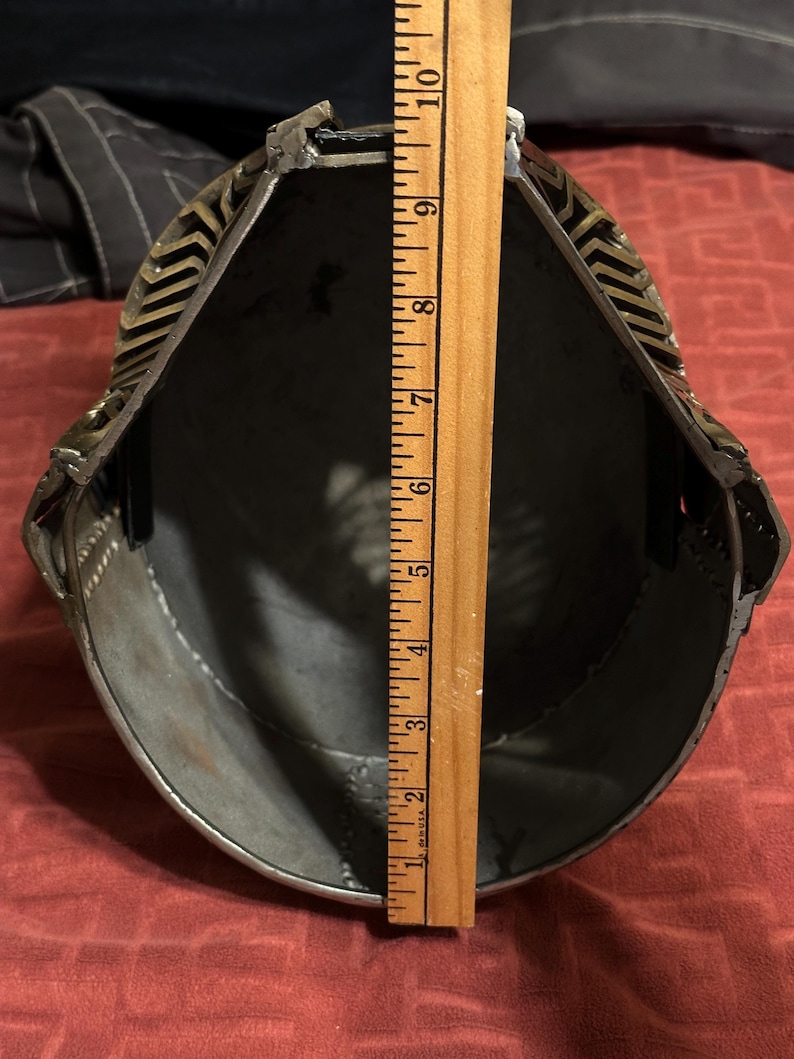May include: A metal helmet viewed from the interior, featuring a dark grey inner surface and a decorative brass-toned exterior trim. A wooden ruler is placed vertically across the opening, showing a measurement of approximately 10 inches in length.