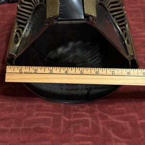 May include: A metallic helmet with a dark finish and gold-toned accents featuring intricate cut-out patterns along the sides. A wooden ruler is positioned across the front opening, showing a width measurement of approximately 8.5 inches.