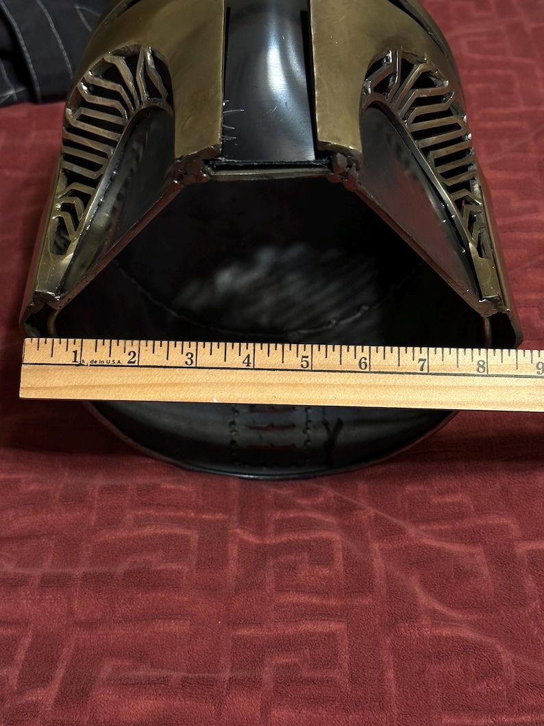 May include: A metallic helmet with a dark finish and gold-toned accents featuring intricate cut-out patterns along the sides. A wooden ruler is positioned across the front opening, showing a width measurement of approximately 8.5 inches.