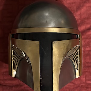 May include: A full-face helmet featuring a smooth, rounded metallic silver dome and a dark black visor. The front is accented with gold-toned metal plating, including a T-shaped faceplate and decorative side vents with intricate line detailing.