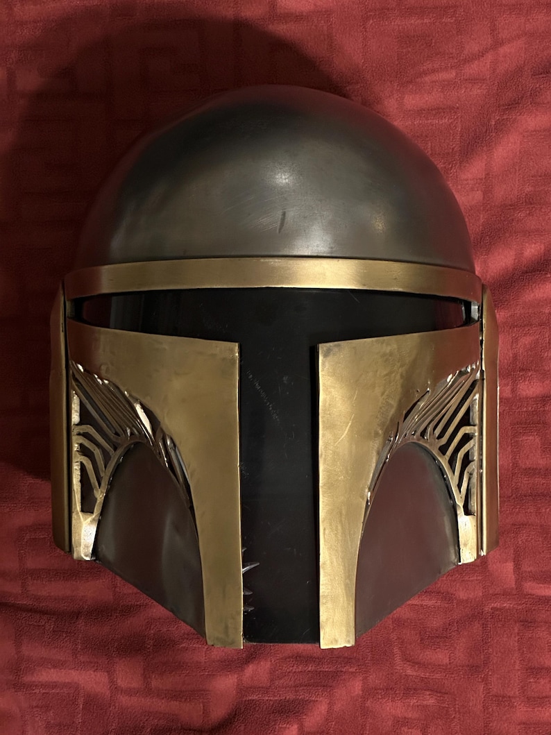 May include: A full-face helmet featuring a smooth, rounded metallic silver dome and a dark black visor. The front is accented with gold-toned metal plating, including a T-shaped faceplate and decorative side vents with intricate line detailing.