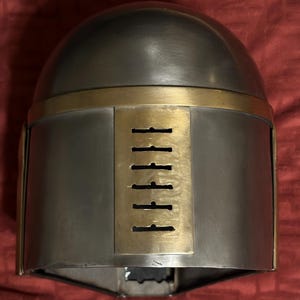 May include: A metallic helmet featuring a smooth, rounded dome and a rectangular brass-colored faceplate. The faceplate includes a vertical row of six horizontal ventilation slits. The helmet has a polished grey finish with brass-colored trim.