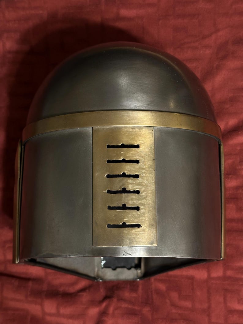 May include: A metallic helmet featuring a smooth, rounded dome and a rectangular brass-colored faceplate. The faceplate includes a vertical row of six horizontal ventilation slits. The helmet has a polished grey finish with brass-colored trim.