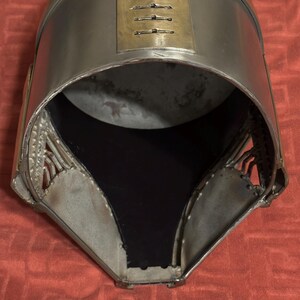 May include: A metallic helmet featuring a smooth, silver-toned finish with a prominent gold-colored band across the top. The front section includes a vertical rectangular plate with six horizontal ventilation slits and a wide, open face opening.