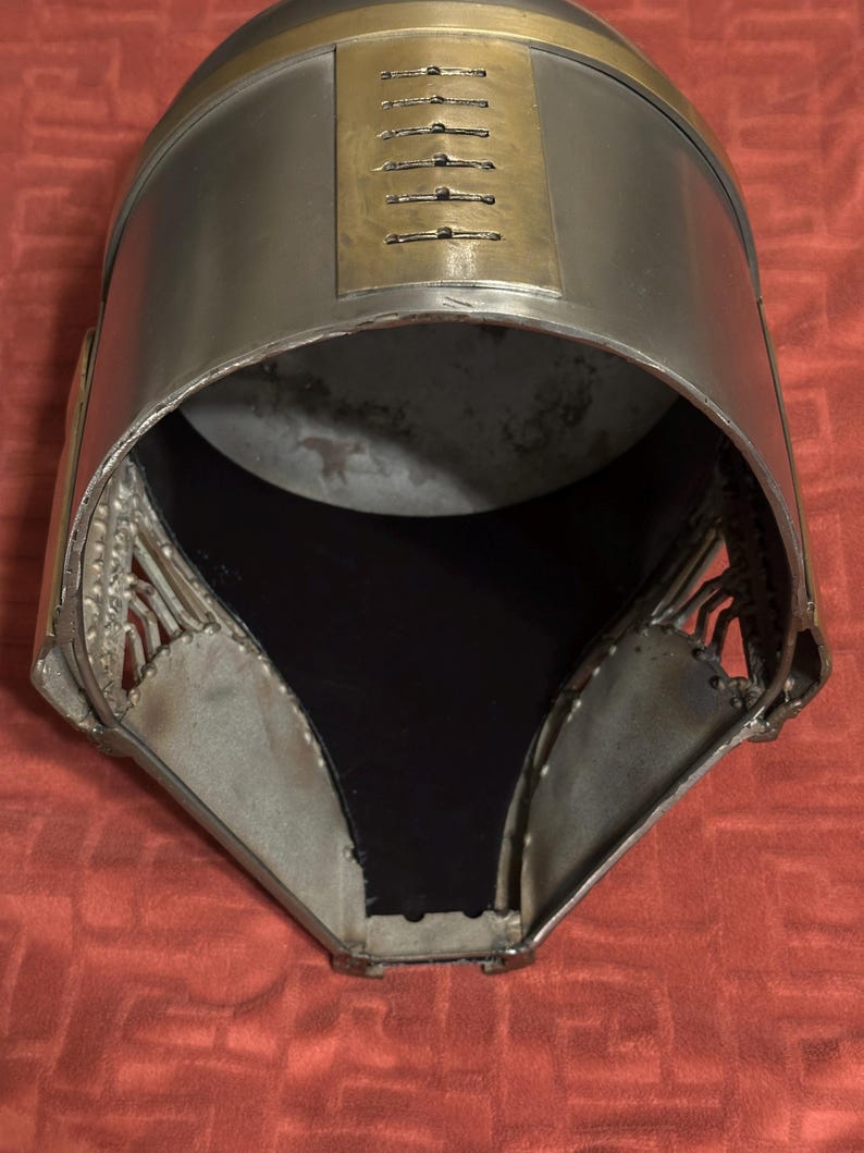 May include: A metallic helmet featuring a smooth, silver-toned finish with a prominent gold-colored band across the top. The front section includes a vertical rectangular plate with six horizontal ventilation slits and a wide, open face opening.