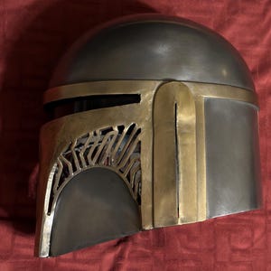 May include: A metallic helmet featuring a smooth, rounded dome and a distinct T-shaped visor design. The helmet is constructed with a combination of dark grey, brushed metal surfaces and contrasting gold-toned metal accents along the faceplate.