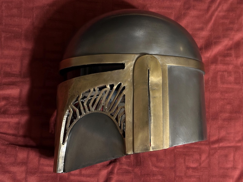 May include: A metallic helmet featuring a smooth, rounded dome and a distinct T-shaped visor design. The helmet is constructed with a combination of dark grey, brushed metal surfaces and contrasting gold-toned metal accents along the faceplate.