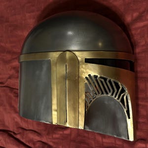 May include: A metallic helmet featuring a smooth, dark gray dome and a contrasting gold-colored faceplate. The faceplate includes a T-shaped visor opening with vertical slats on the right side, showcasing a distinct, angular design.