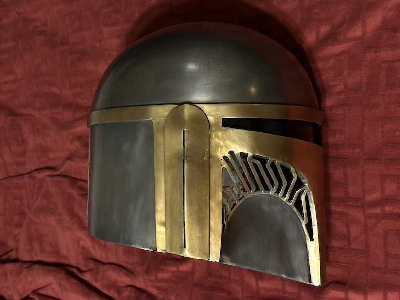 May include: A metallic helmet featuring a smooth, dark gray dome and a contrasting gold-colored faceplate. The faceplate includes a T-shaped visor opening with vertical slats on the right side, showcasing a distinct, angular design.