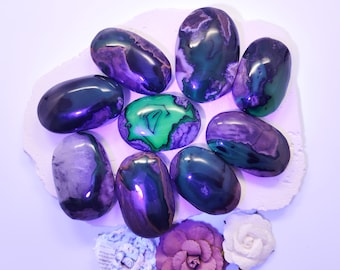 May include: A collection of eight polished agate stones in shades of purple, green, and black. The stones are arranged in a circle on a white surface.
