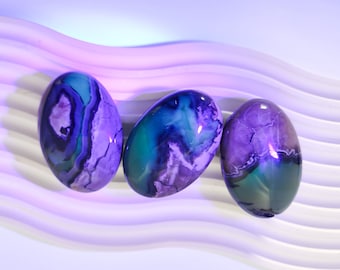 May include: Three oval-shaped agate stones with purple, green, and white swirling patterns. The stones are arranged in a row on a white surface.