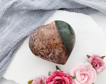 May include: A heart-shaped ceramic sculpture with a brown and green glaze. The sculpture is sitting on a white surface with pink flowers.