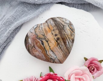 May include: A heart-shaped polished stone with brown, white, and yellow swirling patterns. The stone is on a white surface with a light blue fabric in the background.