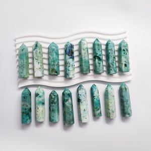 Könnte beinhalten: Eine Sammlung von 18 grünen und weißen Kristallspitzen, die in zwei Reihen auf einer weißen, wellenförmigen Oberfläche angeordnet sind. Die Spitzen sind alle ungefähr gleich groß und geformt und haben eine natürliche, raue Textur.