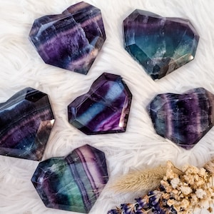 May include: Six heart-shaped fluorite crystals in shades of purple, green, and black. The crystals are arranged on a white fluffy surface.
