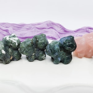 May include: Five small carved animal figurines made of different types of stone. The figurines are arranged in a row on a white surface. From left to right, the figurines are made of green moss agate, green moss agate, blue moss agate, pink rose quartz, and black obsidian.
