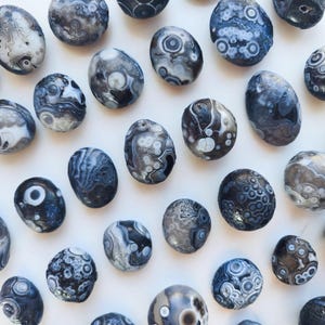 May include: A collection of polished orbicular agate stones featuring intricate circular patterns in shades of charcoal, slate gray, and white. Each stone displays unique concentric banding and eye-like formations scattered across the surface.