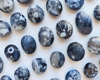 May include: A collection of polished orbicular agate stones featuring intricate circular patterns in shades of charcoal, slate gray, and white. Each stone displays unique concentric banding and eye-like formations scattered across the surface.