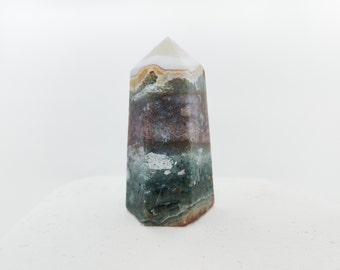 Puede incluir: Una punta de cristal de ágata con bandas verdes, marrones y blancas, también conocida como varita de cristal, es una piedra natural con una punta puntiaguda. El cristal está sobre una superficie blanca.