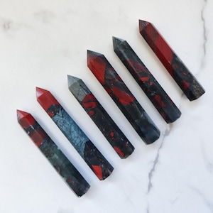 May include: Six polished stone wands arranged in a diagonal row. Each wand features a hexagonal prism shape with a pointed tip. The stones display a mix of deep black, dark gray, and vibrant red colors with unique natural patterns and textures.
