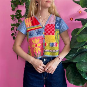 Può includere: Un cardigan all'uncinetto a maniche corte caratterizzato da un vivace design patchwork con quadrati rossi, gialli, rosa, verde lime e blu. Il capo presenta uno scollo a V, una chiusura frontale con bottoni multicolori e motivi come un fiore giallo.