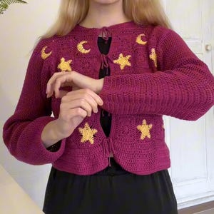 May include: A magenta crochet cardigan with yellow star and crescent moon appliqués. The long-sleeved sweater has a round neckline and a tie closure. The celestial-themed garment is worn over a black top.