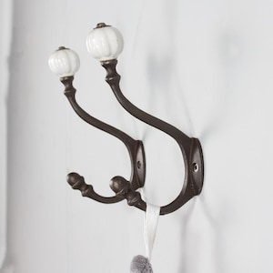 May include: Two antique-style wall hooks. The hooks are dark brown with white, pumpkin-shaped ceramic knobs. The hooks are mounted on a white wall. A white ribbon is hanging from one of the hooks.