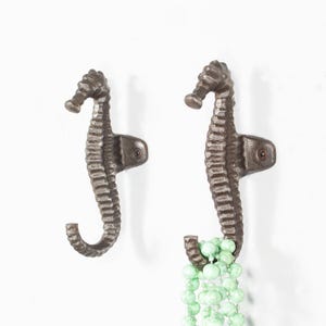 May include: Two dark brown, textured metal wall hooks shaped like seahorses. Each hook features a mounting bracket with a single screw hole. The right hook holds a strand of light green, round beads draped over its curved tail section.