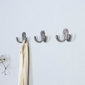 May include: Three silver double hooks mounted on a white wall. A white scarf is hanging on one of the hooks.