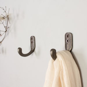 May include: Two antique-style metal hooks mounted on a white wall. One hook has a white scarf draped over it.