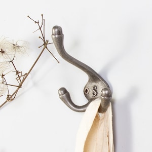 May include: A silver metal hook with three prongs. The hook is mounted on a white wall and has a small letter 'S' on the back.