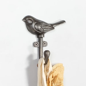 May include: A dark grey metal wall hook featuring a sculpted bird figure at the top. The hook has a vertical mounting plate with two visible screws and a curved lower prong that holds a light-coloured fabric item with a textured, ruffled edge.