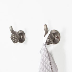 May include: Two cast iron wall hooks shaped like thumbs up. The hooks are dark grey and mounted on a white wall. One hook holds a grey and white patterned towel. The thumbs up design adds a unique touch to any space.