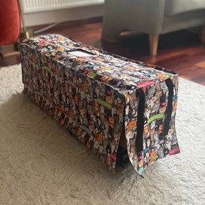 May include: A rectangular cat bed with a colorful pattern of various cat breeds. The bed has a rectangular opening on top and black handles on the sides. The cat bed is placed on a white rug.