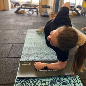 May include: A woman is doing a forearm plank on a black and teal yoga mat with a mandala pattern. The mat has the text "You are worth it" and "Love your core" printed on it.