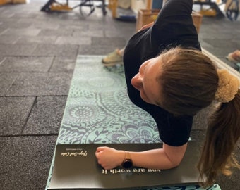 May include: A woman is doing a forearm plank on a black and teal yoga mat with a mandala pattern. The mat has the text "You are worth it" and "Love your core" printed on it.