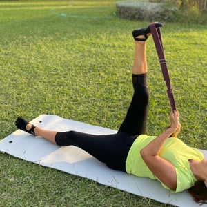 May include: A person stretches their leg using a purple yoga strap while lying on a gray yoga mat. They are wearing black leggings and a bright yellow shirt.