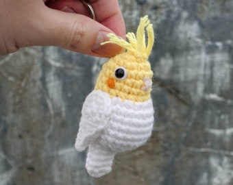 Può includere: Un portachiavi a uncinetto a forma di cacatua ninfa giallo e bianco con occhi neri e un piccolo becco arancione.
