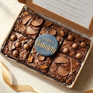 May include: A cardboard box containing six square chocolate brownies topped with various chocolate candies and orange slices. A round, dark blue disc with the gold embossed text "HAPPY Birthday" is placed in the centre of the brownies.