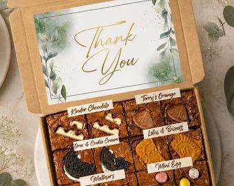 May include: An open cardboard box filled with brownies, each topped with different treats like Kinder Chocolate and Lotus Biscoff. The box lid has a card that says "Thank You". The brownies are presented on a marble surface, with a cake visible.