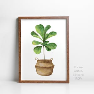 May include: A cross stitch pattern PDF featuring a watercolor-style illustration of a green fiddle leaf fig plant in a tan woven basket with handles, displayed inside a brown wooden frame. The text Cross stitch pattern (PDF) is visible.