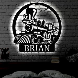 Peut inclure: Silhouette noire en métal d'une locomotive avec le nom "BRIAN" en dessous. La silhouette est montée sur un mur en briques et éclairée par une lumière blanche.