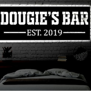May include: Black and white sign with the text "Dougie's Bar Est. 2019" on a brick wall background.