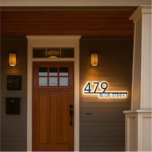 May include: Illuminated address sign displaying "479 BLUE STREET" in a modern font. The sign is mounted on a gray exterior wall, with a wooden door and porch lights visible. The sign is lit with a warm, inviting glow.