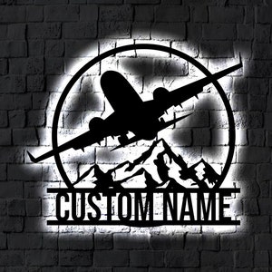 May include: Black metal silhouette of an aeroplane flying over mountains, inside a circle, with the text "CUSTOM NAME" below.